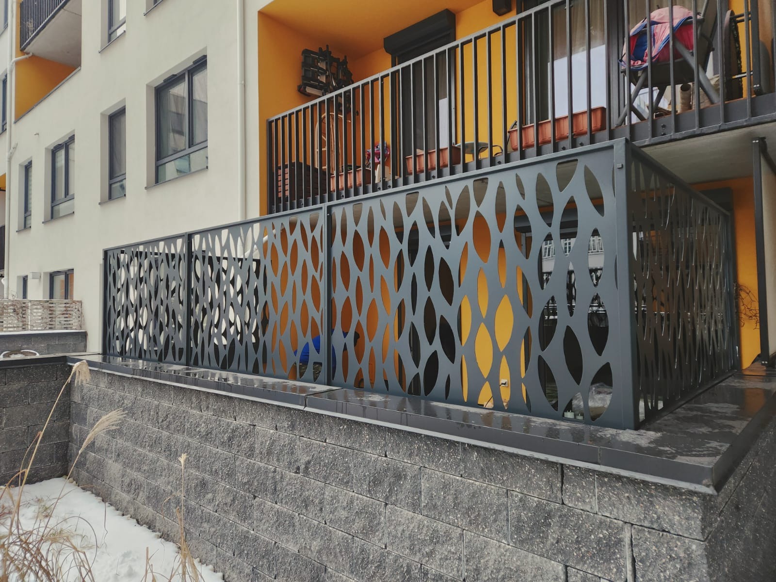 Nowoczesna, ażurowa balustrada balkonowa z motywem liści, wykonana z szarego metalu. Tło stanowi budynek z żółtym balkonem. Widoczny fragment murku z kamienia.