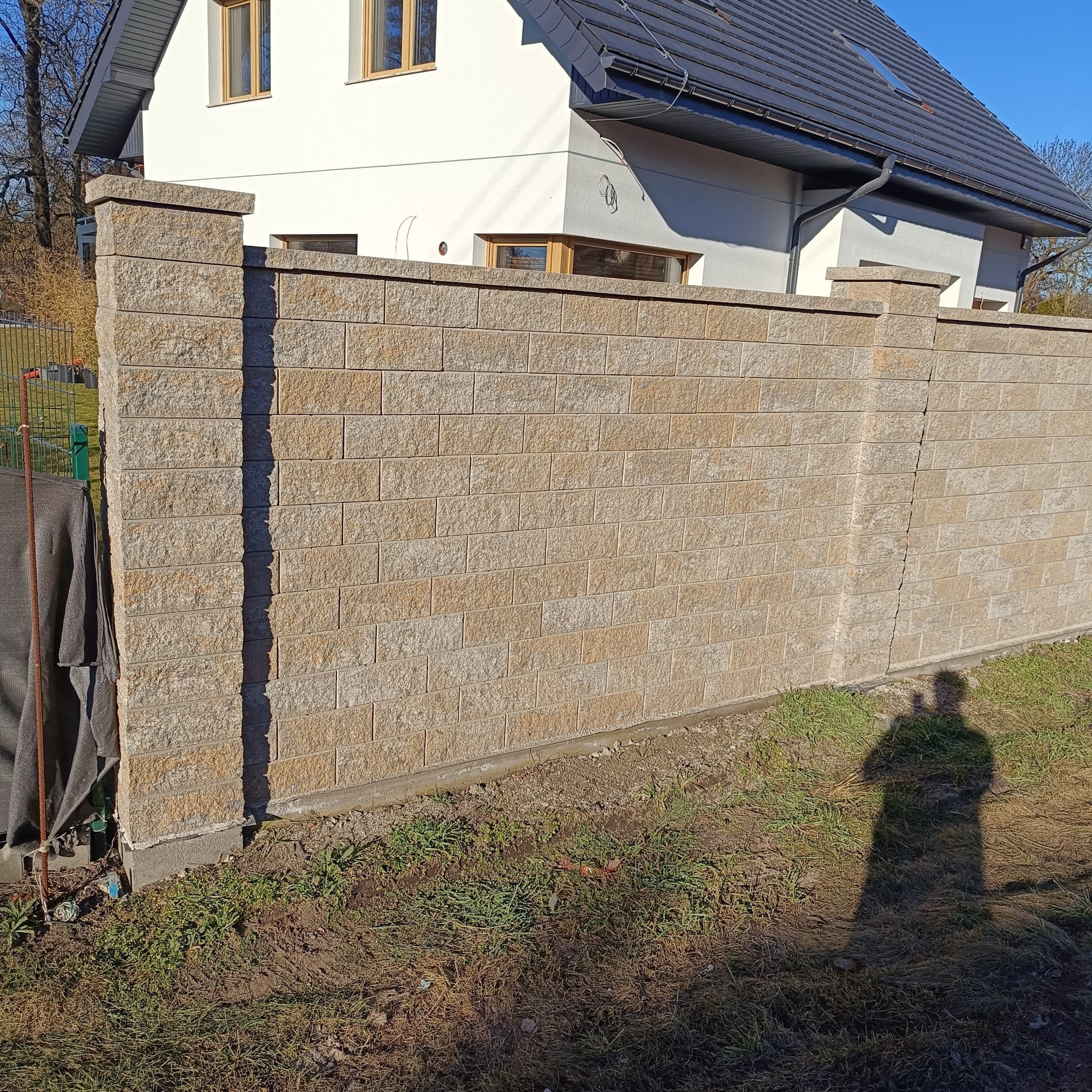 Nowoczesny, beżowy mur z bloczków betonowych, oddzielający posesję od ulicy. W tle biały dom z ciemnym dachem i zielony trawnik. Widoczny cień osoby robiącej zdjęcie.