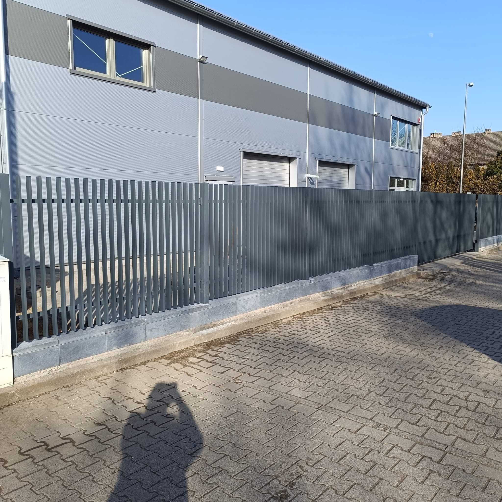 Nowoczesne ogrodzenie metalowe w Zawoni, z pionowymi sztachetami i podmurówką z szarej kostki, na tle budynku przemysłowego w odcieniach szarości.