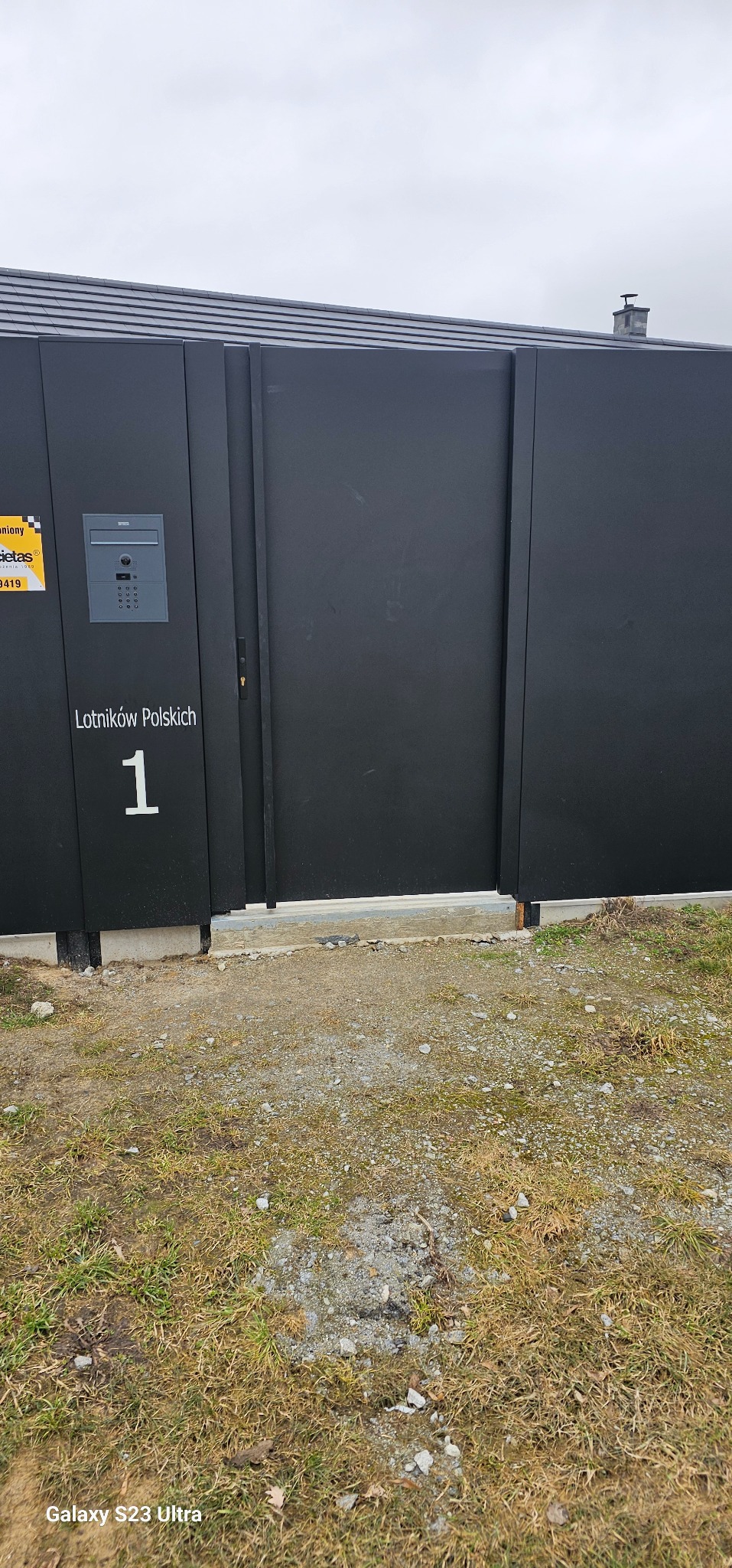 Nowoczesna, przesuwna brama w kolorze antracytu z numerem posesji i domofonem, zamontowana przy posesji z widocznym fragmentem budynku w tle.