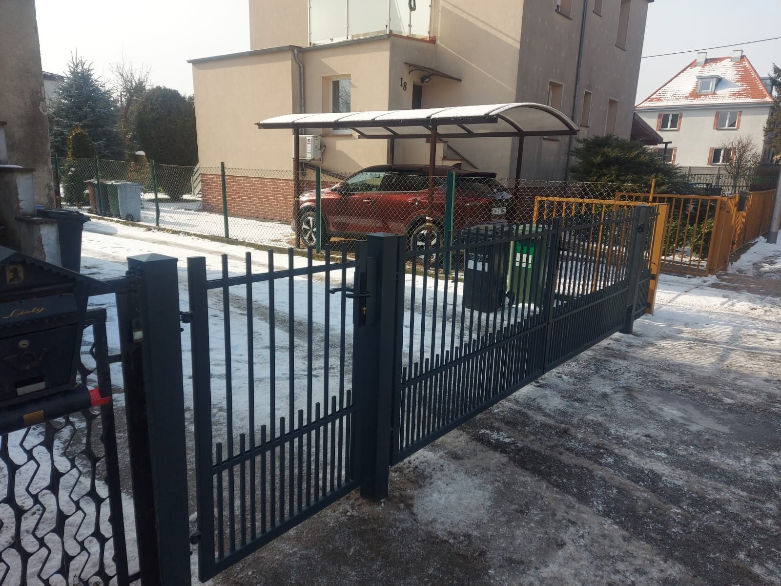 Nowoczesna, grafitowa brama wjazdowa z pionowymi szczebelkami, z widocznym fragmentem posesji i samochodem pod wiatą. Zima, lekko ośnieżona nawierzchnia.