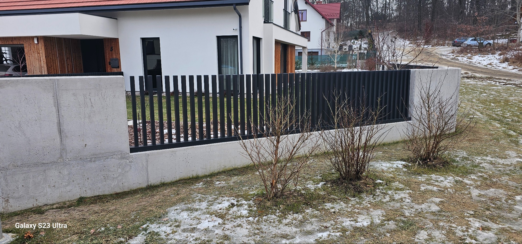 Nowoczesne ogrodzenie metalowe z betonowymi słupkami, pionowe panele w kolorze antracytu, na tle domu z elementami drewna i ogrodu z resztkami śniegu.