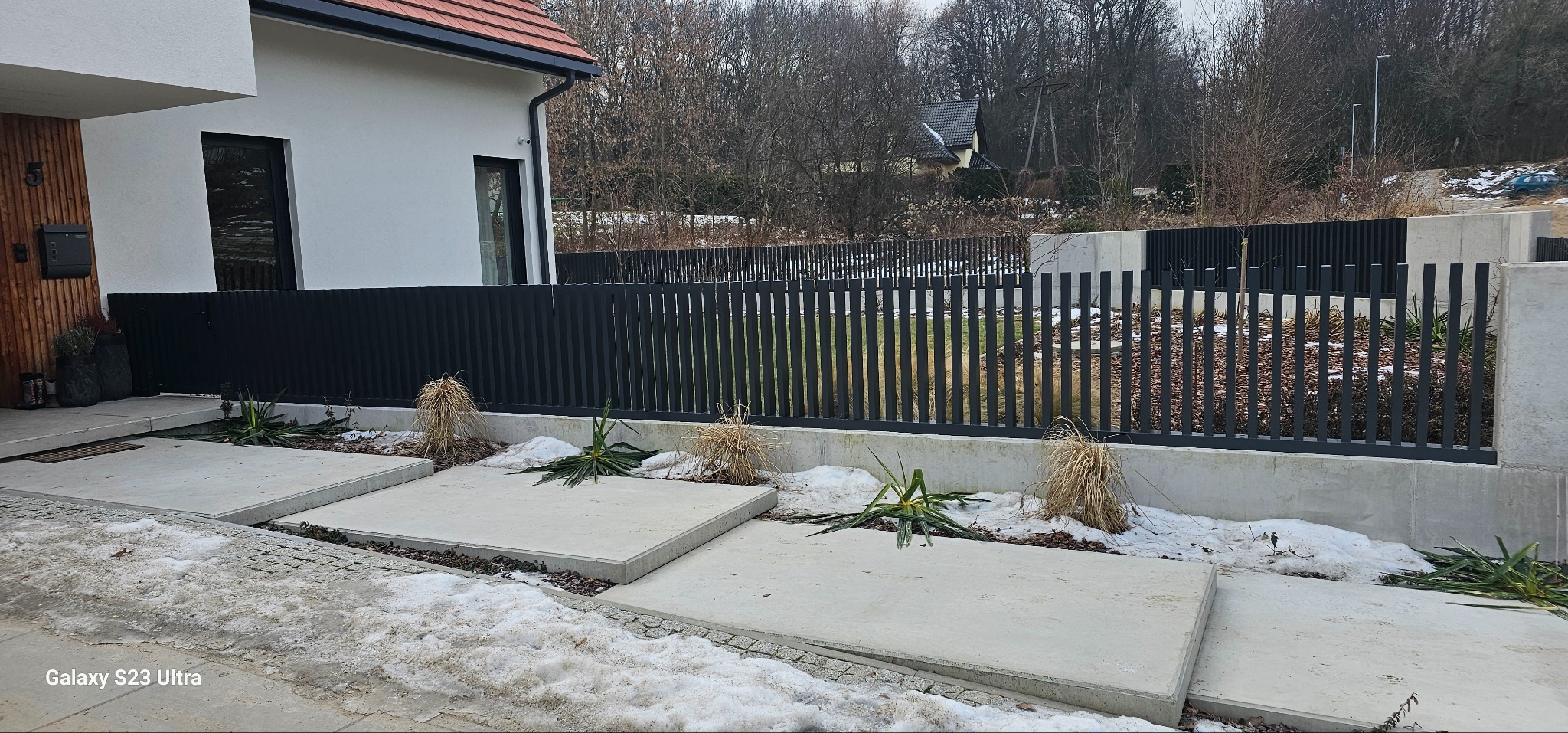 Nowoczesne ogrodzenie metalowe w kolorze antracytowym, z pionowymi sztachetami, osadzone na betonowej podmurówce. Widoczne elementy architektury domu i zimowy krajobraz w tle.