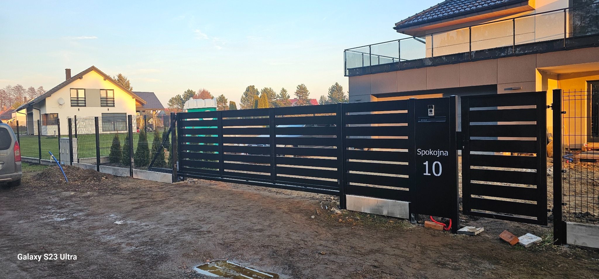 Nowoczesna, przesuwna brama wjazdowa z poziomymi panelami, numer domu 'Spokojna 10', skrzynka na listy, widok na dom i ogrodzenie panelowe w tle, pochmurne niebo.