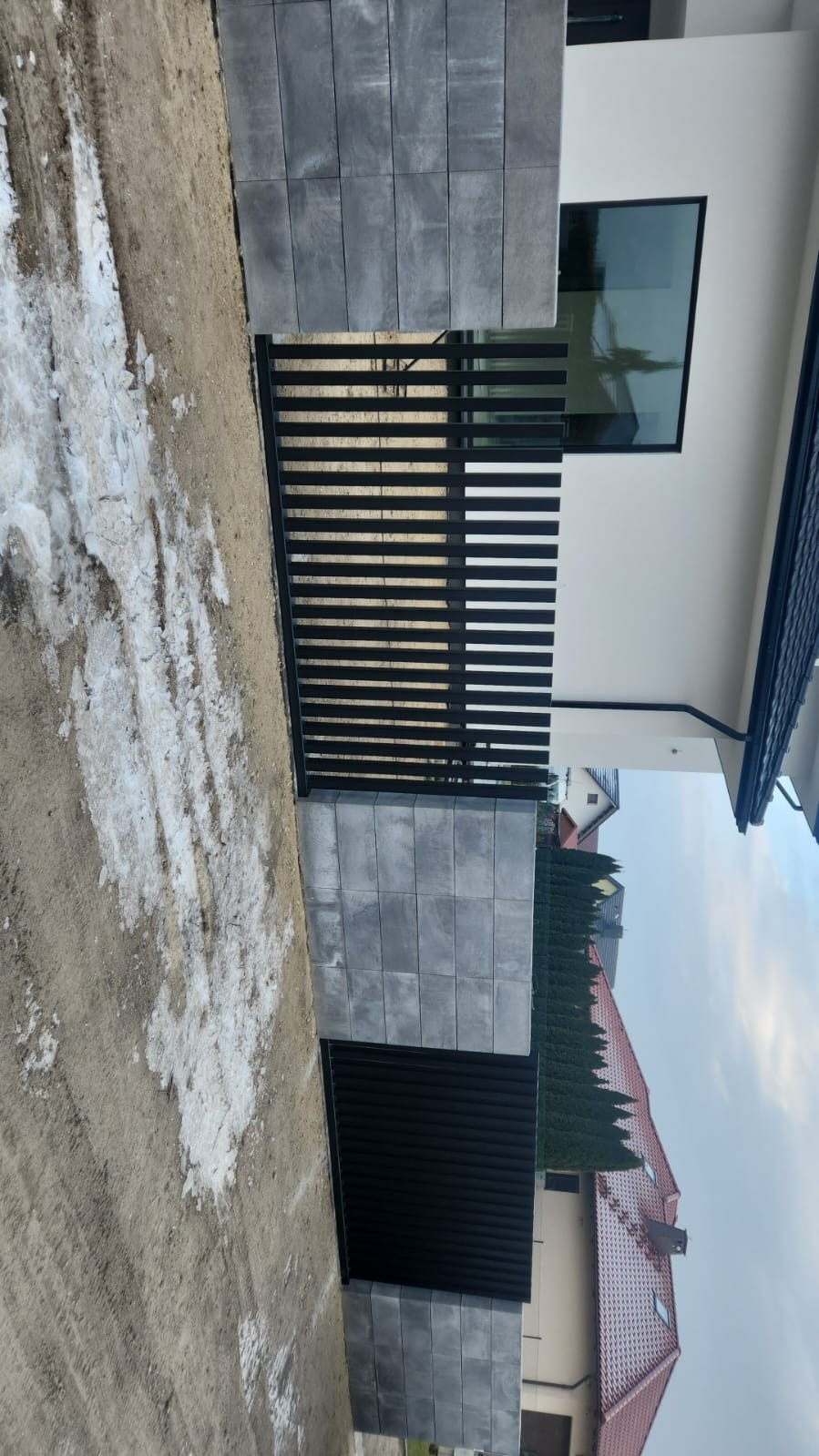 Nowoczesna brama wjazdowa z poziomymi, czarnymi przęsłami, osadzona na szarych, betonowych słupkach. Widok na dom z oknem i fragment sąsiedniej posesji.