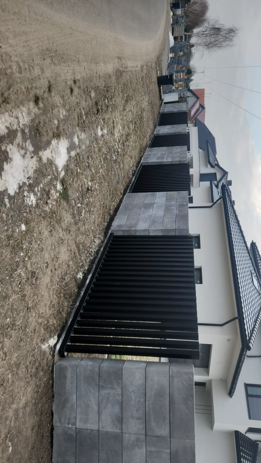 Nowoczesne ogrodzenie z czarnych, poziomych paneli i szarych, betonowych słupków, oddzielające posesję od drogi. Widok z boku, w tle dom z ciemną dachówką.