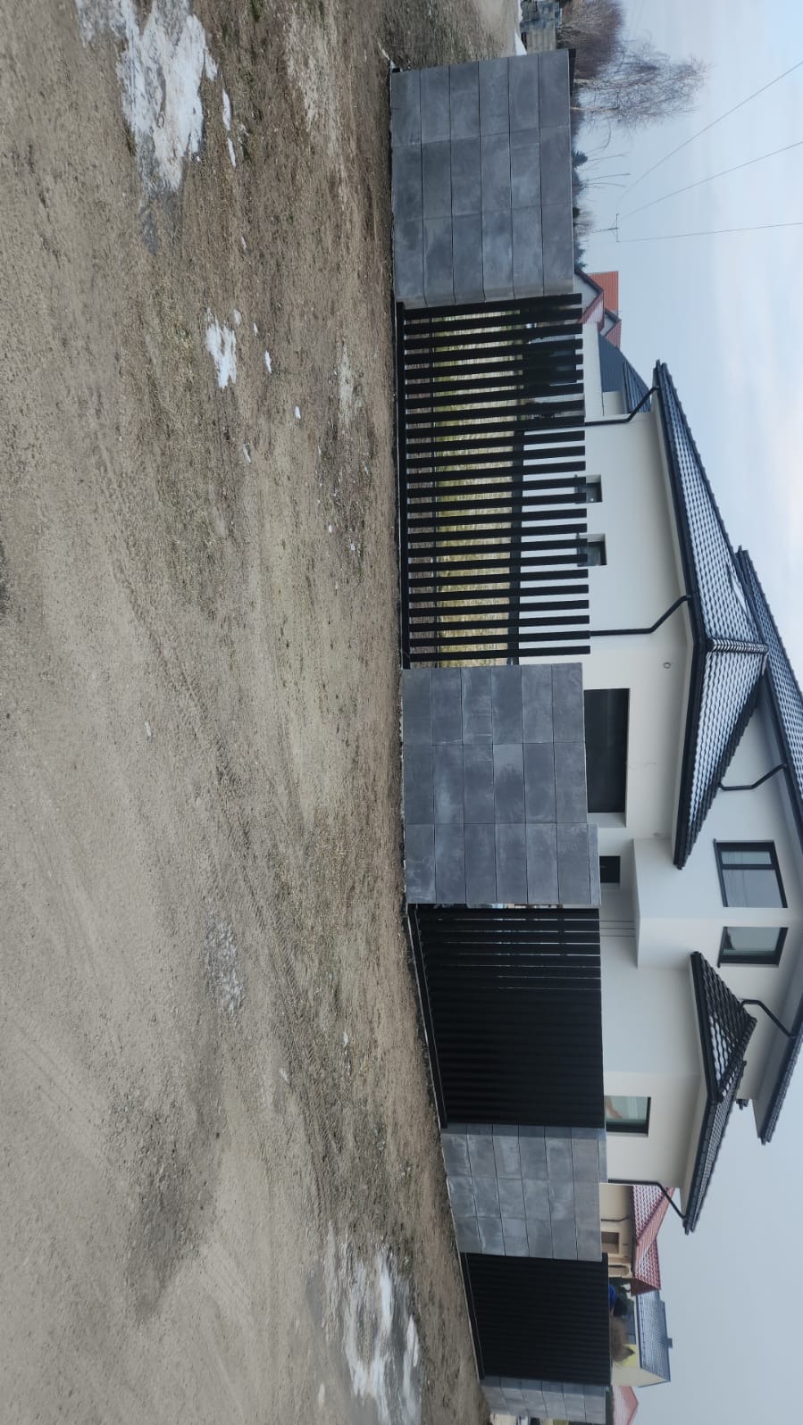 Nowoczesna, czarna brama posesyjna z szarymi, betonowymi słupkami na tle białego domu z ciemną dachówką. Minimalistyczny design, surowy krajobraz.