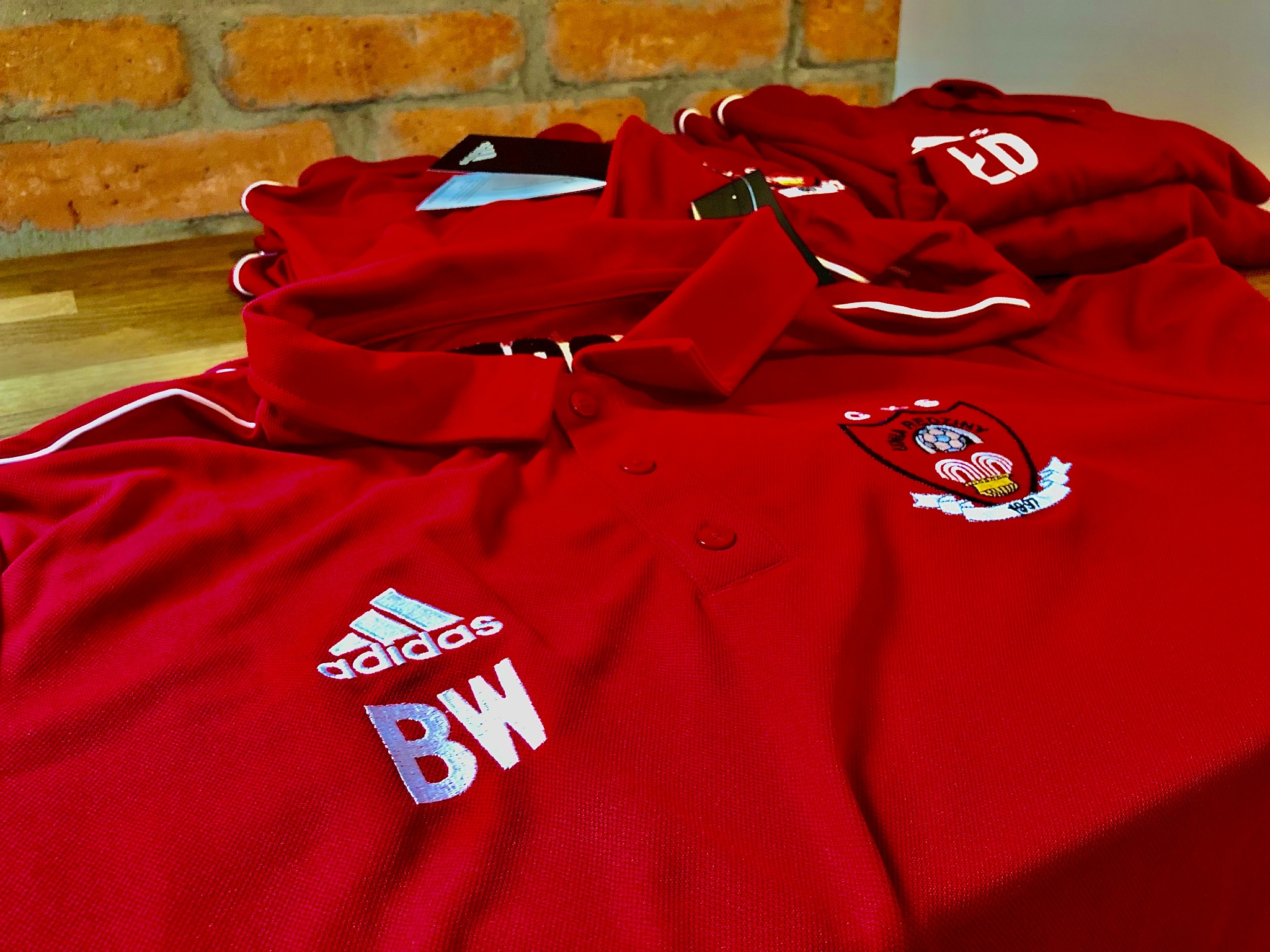Stos czerwonych koszulek polo z haftowanymi herbami i inicjałami, leżący na drewnianym blacie na tle ceglanej ściany.