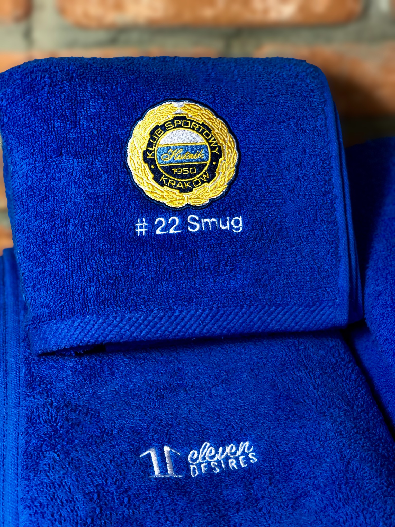 Stos niebieskich ręczników frotte z wyhaftowanym logo klubu sportowego z Krakowa, numerem #22 i nazwą '11 eleven DESIRES'.