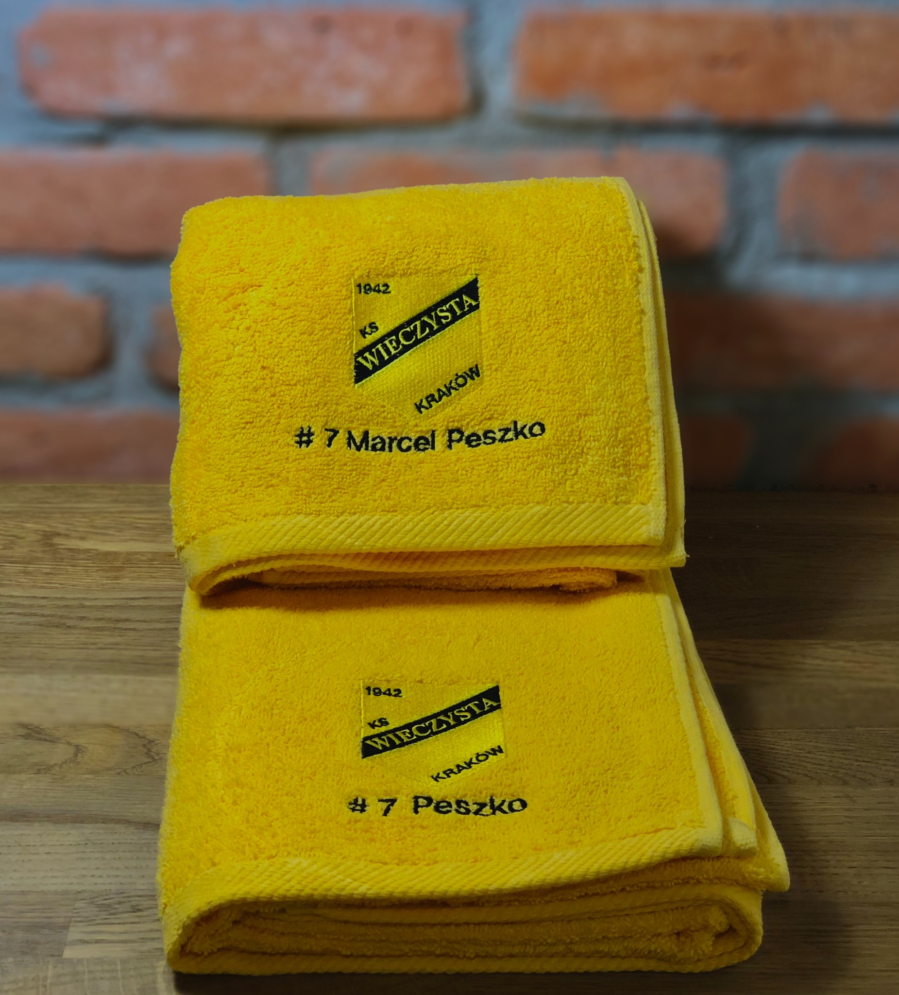 Dwa żółte ręczniki frotte, jeden na drugim, z czarnym haftem: logo KS Wieczysta Kraków z rokiem 1942 i nazwiskiem '#7 Marcel Peszko' na górnym i '#7 Peszko' na dolnym, na tle ceglanej ściany...