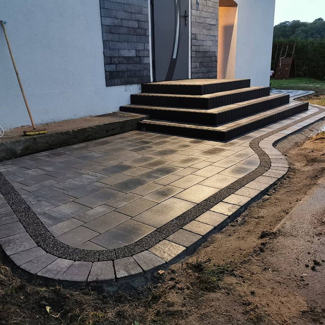 Ułożona szara kostka brukowa przed domem z ciemnymi schodami wejściowymi, obrzeża z drobnego grysu, widoczny fragment elewacji i drzwi wejściowe.