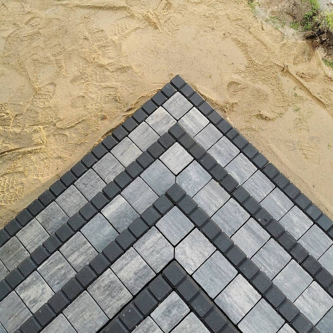 Ułożona, szara kostka brukowa w geometryczny wzór z ciemnoszarą obwódką, widoczna na piasku z odciskami butów.