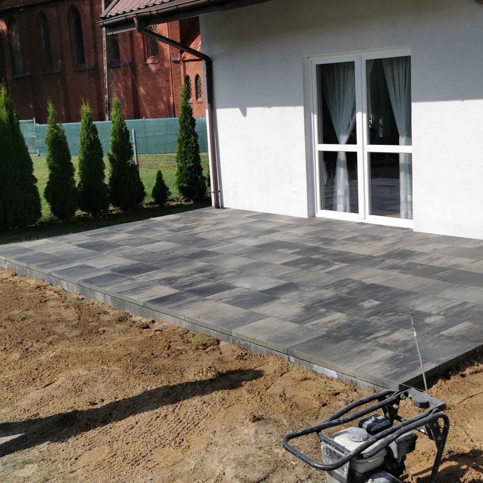 Nowo ułożony, szaro-czarny taras z kostki brukowej w kratkę, przylegający do białego budynku z oknem, z ubijarką wibracyjną na pierwszym planie i rzędem tuj w tle.