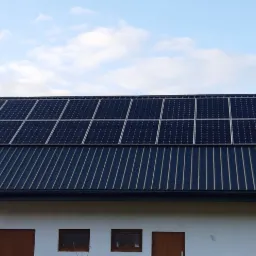 Instalacja paneli słonecznych na dachu budynku gospodarczego z blachy trapezowej, widoczne ciemne panele w srebrnych ramach pod błękitnym niebem z chmurami.