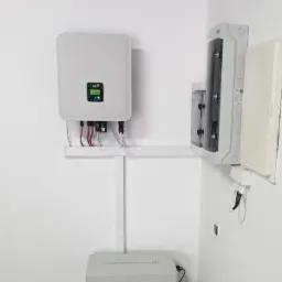 Biała ściana z zamontowanym inwerterem, akumulatorem energii FOX ESS i skrzynką elektryczną. Widoczne przewody i elementy montażowe.