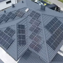 Widok z drona na dach domu pokryty panelami słonecznymi w różnych konfiguracjach, z elementami krajobrazu w tle.