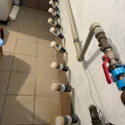 Iniekcja krystaliczna w piwnicy: rząd nawierconych otworów z aplikatorami preparatu do osuszania murów, widoczne rury instalacyjne i płytki podłogowe.