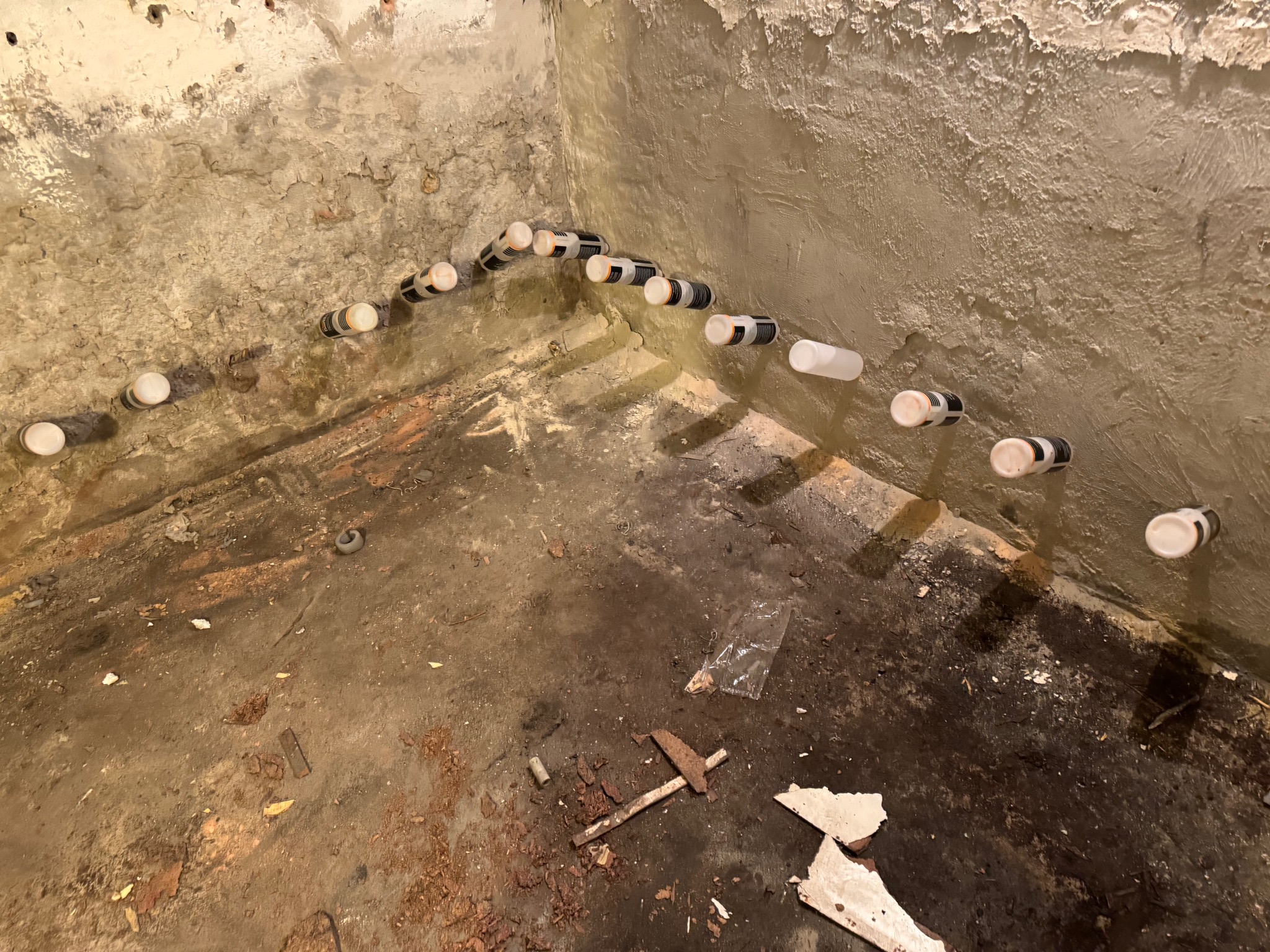 Piwnica w trakcie osuszania: widoczne nawiercone otwory w ścianie z aplikatorami preparatu, wilgotne ślady na podłodze, fragmenty gruzu i starego narzędzia na posadzce.