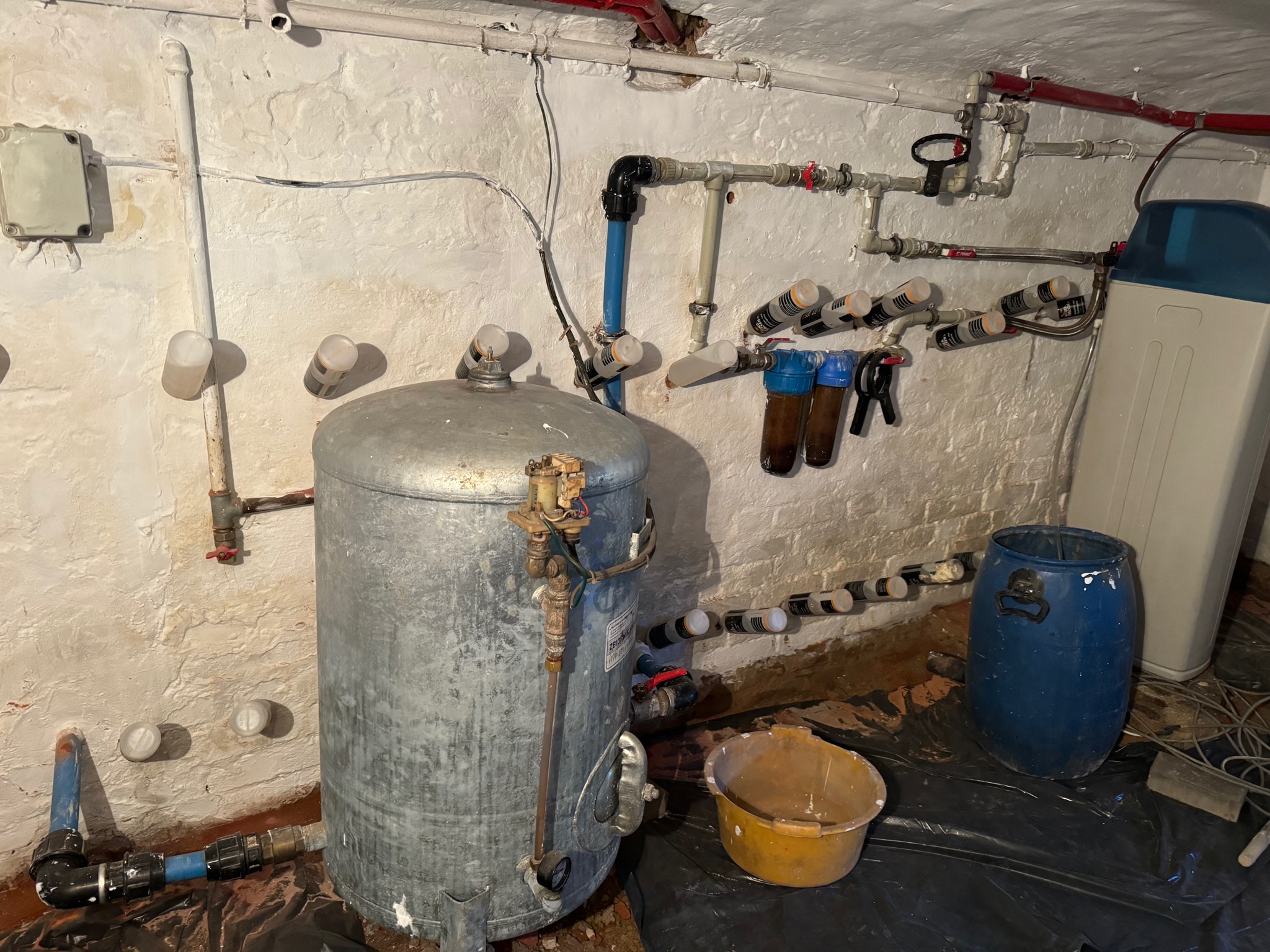 Metalowy zbiornik ciśnieniowy w piwnicy z instalacją wodną, widoczne filtry wody, rury i plastikowy pojemnik na czarnej folii ochronnej.