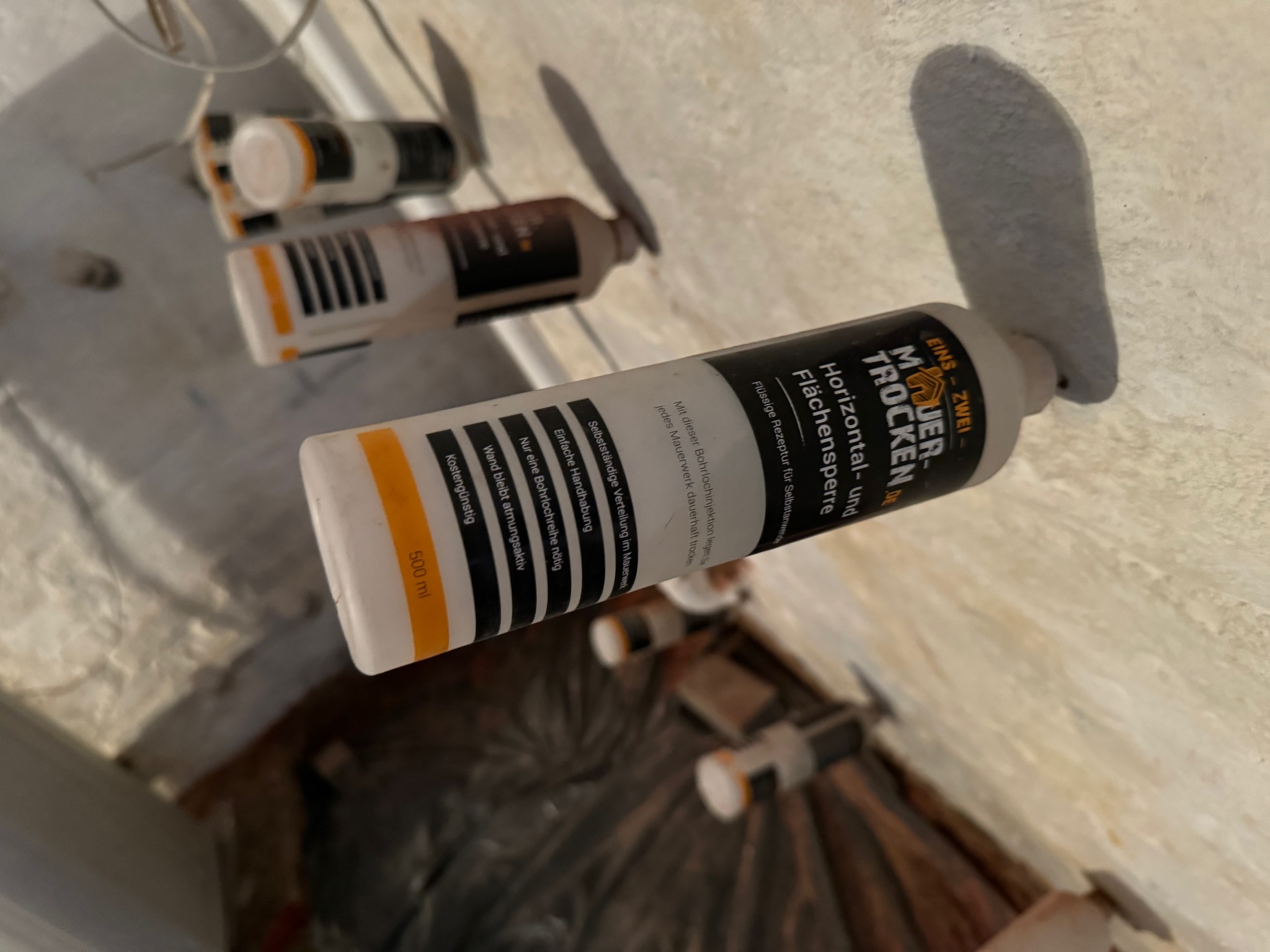 Pojemniki z preparatem do iniekcji w murze, widoczne nawiercone otwory w ścianie, czarna folia zabezpieczająca podłoże.