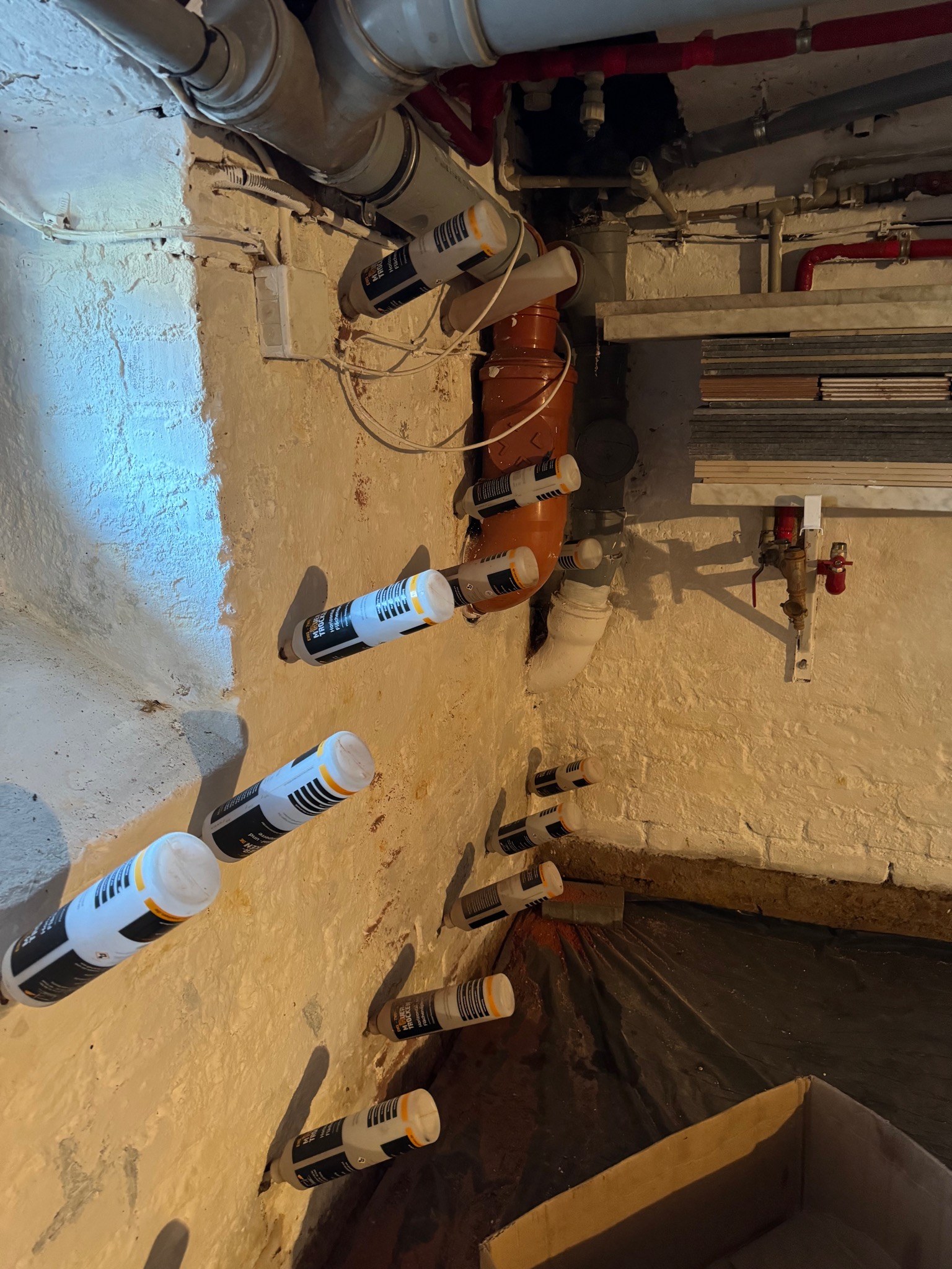 Piwnica z widocznymi nawiertami i aplikacjami preparatu do iniekcji w ścianę w celu osuszenia, widoczne rury instalacji wodnej pod sufitem.