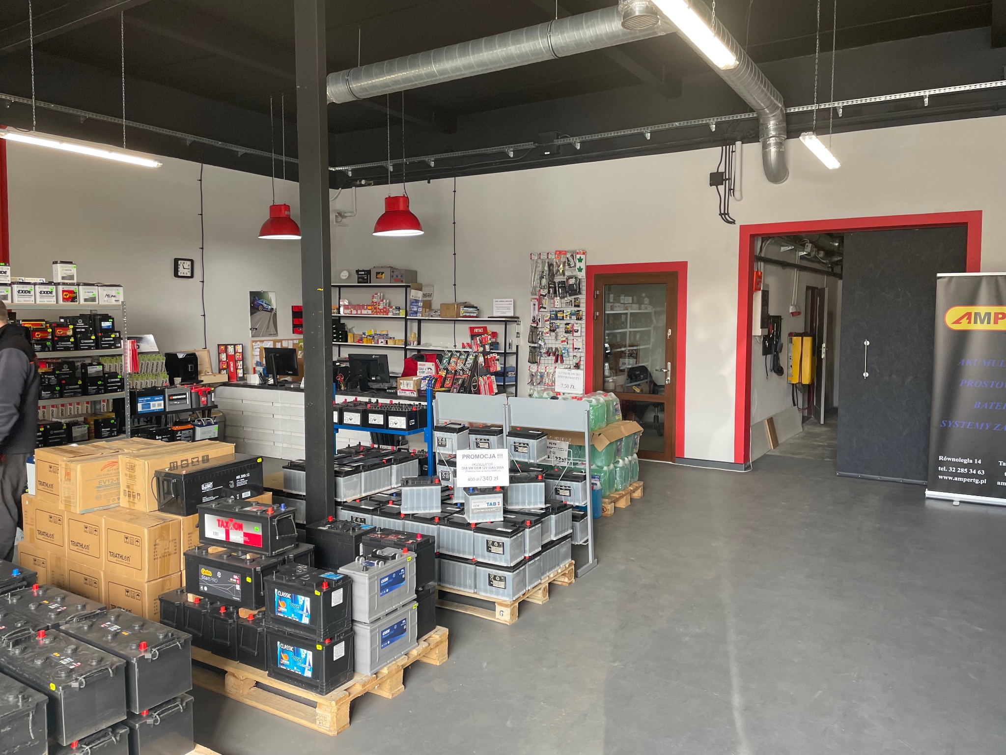 Wnętrze sklepu z częściami samochodowymi po remoncie, widoczne regały z akumulatorami, oświetlenie przemysłowe i świeżo pomalowane ściany w kolorze szarym.