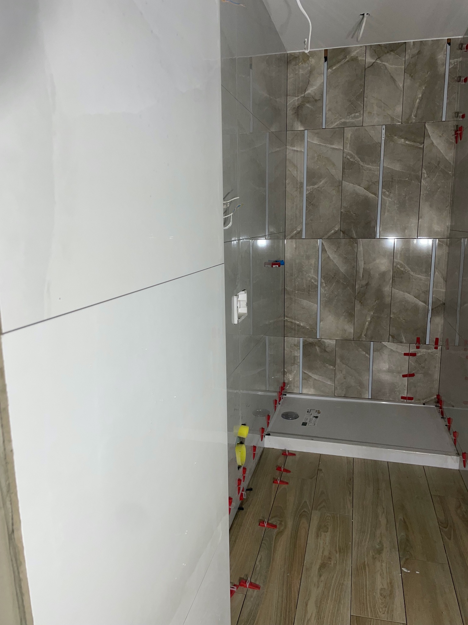 Montaż płytek w łazience: częściowo wykończona ściana z płytek imitujących marmur, biała ściana z połyskiem, brodzik i podłoga z płytek drewnopodobnych, widoczne poziomice i system poziomowania...