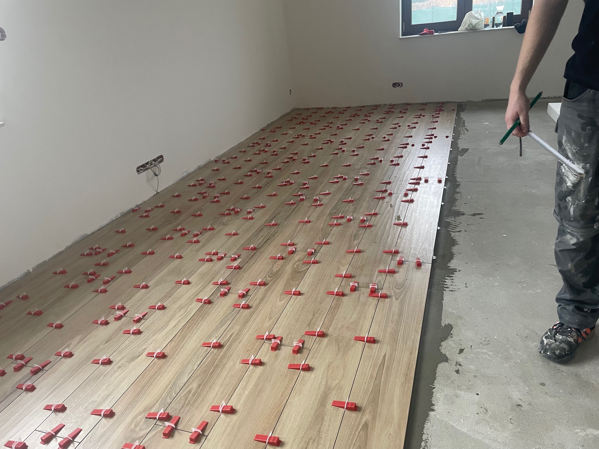 Układanie podłogi z płytek imitujących drewno. Widoczne czerwone kliny dystansowe. W tle ściana z widocznymi puszkami elektrycznymi i fragment okna. Pracownik w ubrudzonych spodniach trzyma narzędzia.