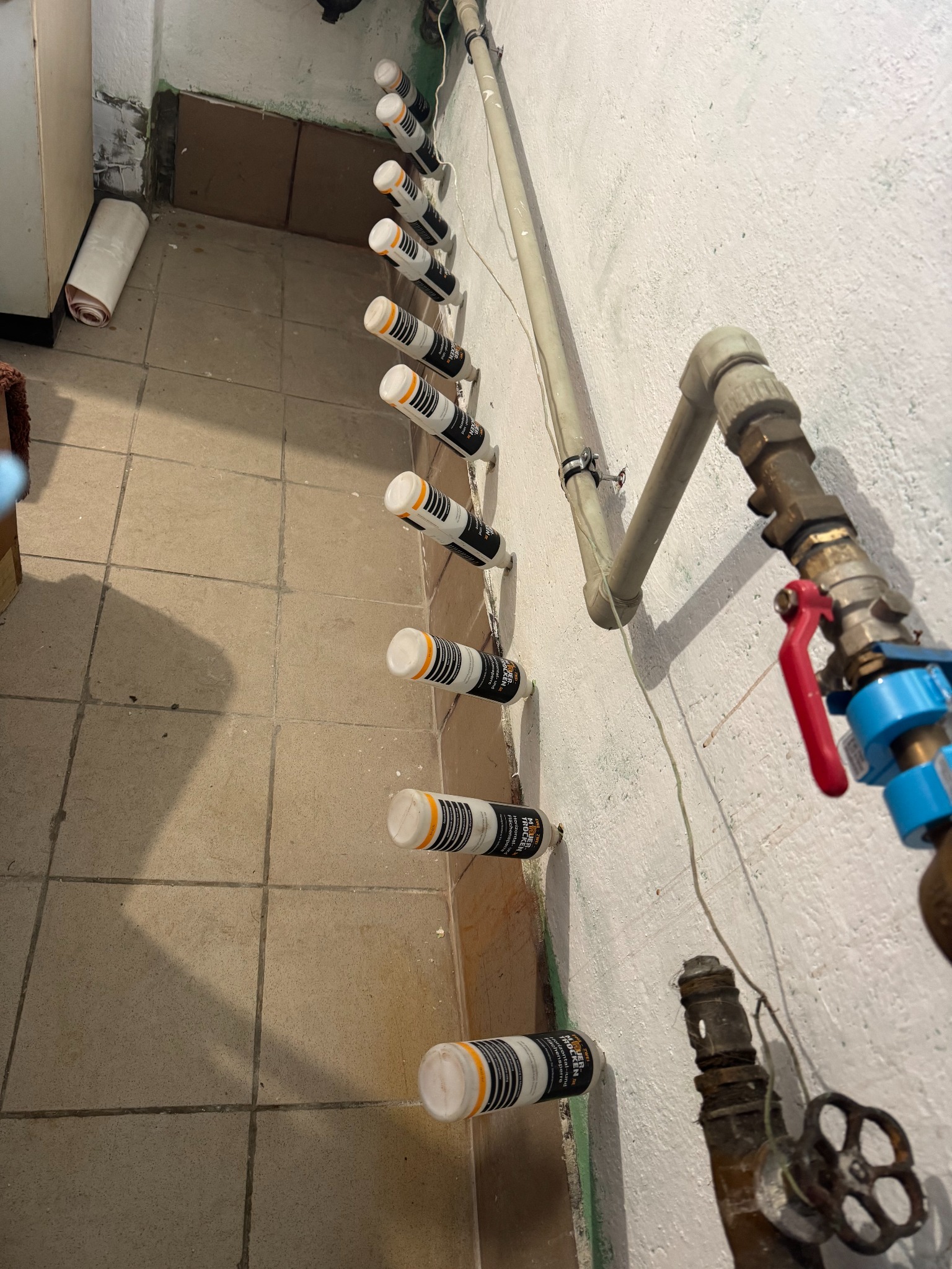 Iniekcja krystaliczna w piwnicy: rząd nawierconych otworów z aplikatorami preparatu do osuszania murów, widoczne rury instalacyjne i płytki podłogowe.