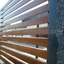 Fragment nowoczesnego ogrodzenia z poziomych, lakierowanych desek w odcieniach brązu, zamocowanych do metalowego słupka, z widocznym fragmentem domu w tle.