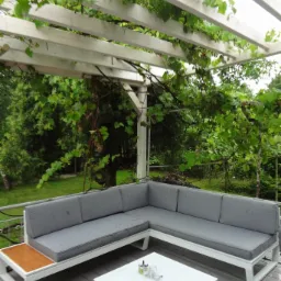 Biała drewniana pergola z pnączami winogron nad szarym narożnikiem ogrodowym i białym stolikiem kawowym na tarasie z desek kompozytowych.