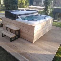Zabudowana wanna SPA z drewnianymi schodkami na tarasie z desek. Widoczny dom w tle.