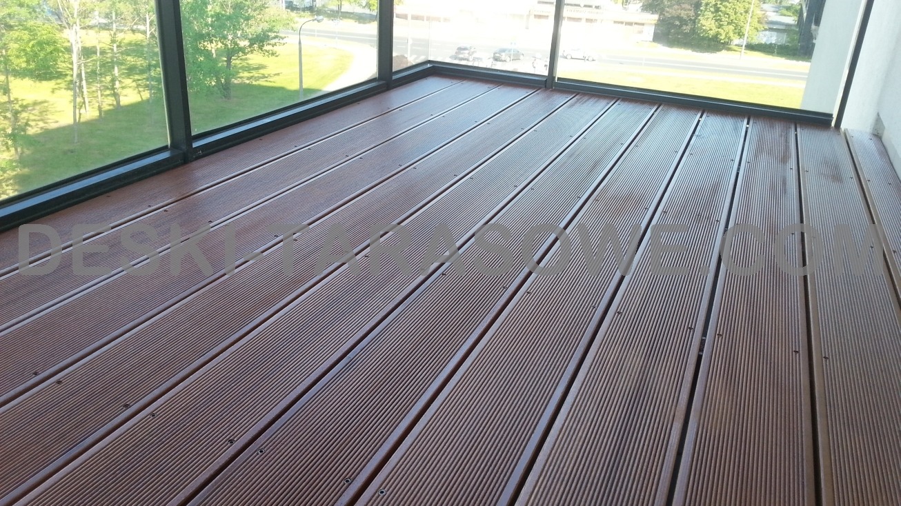 Gładka, brązowa podłoga z desek kompozytowych na balkonie z widokiem na zieleń miejską za oknami.