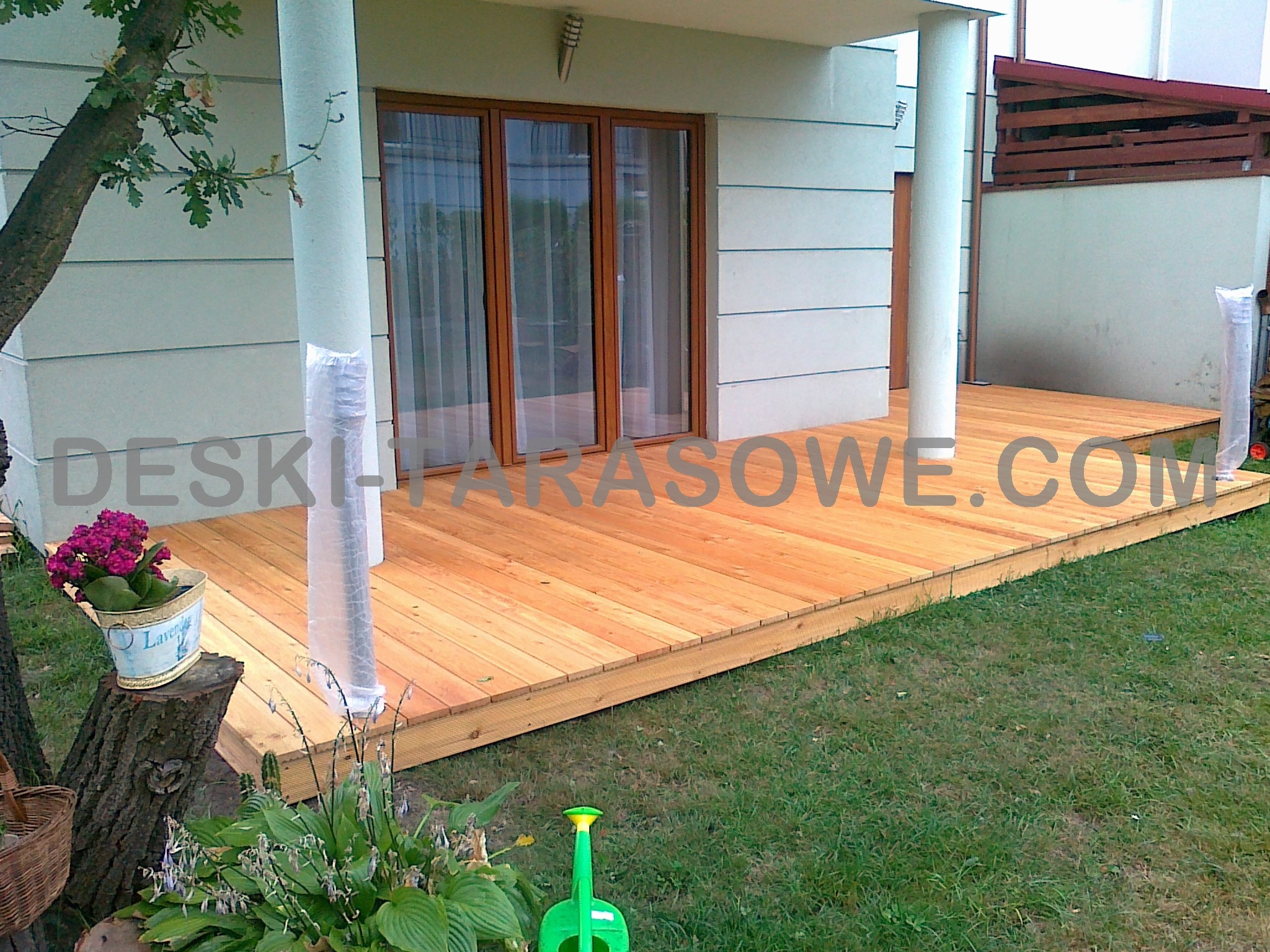 Nowy taras z jasnego drewna przylegający do domu, z kwiatami w ozdobnym wiaderku na pniu drzewa i zielonym konewką na trawniku.