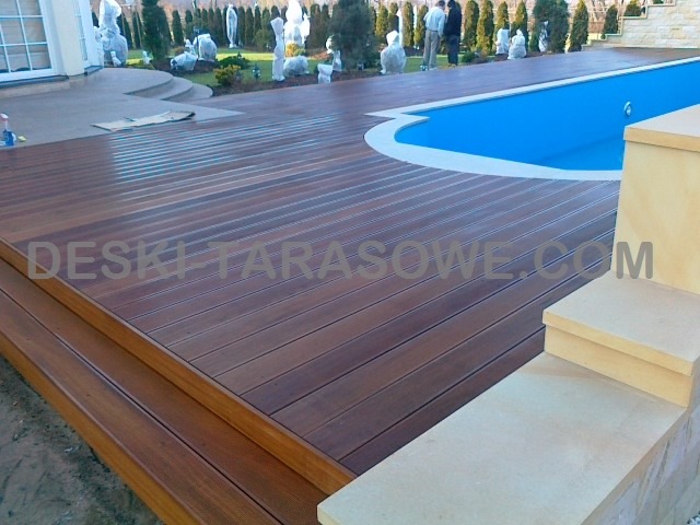 Nowoczesny taras drewniany z ciemnego drewna, przylegający do basenu z jasnoniebieską wodą, schody z jasnego kamienia prowadzące na taras, w tle częściowo zasłonięte rośliny i drzewa.