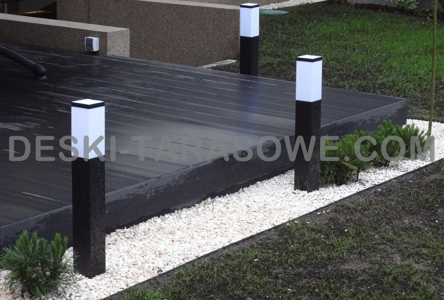 Nowoczesny taras z ciemnych desek kompozytowych, oświetlony trzema lampami stojącymi z białymi kloszami, otoczony białym żwirem i zielenią trawnika.
