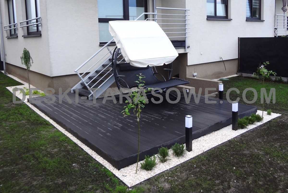 Czarny taras z desek, obrzeże z białego kamienia, oświetlenie w postaci niskich lamp, huśtawka z baldachimem i schody do budynku w tle.