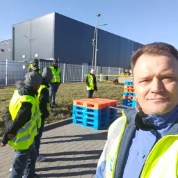 Grupa os&oacute;b w kamizelkach odblaskowych stoi na zewnątrz budynku przemysłowego, obok palet. Na pierwszym planie mężczyzna w kamizelce odblaskowej i niebieskiej kurtce.