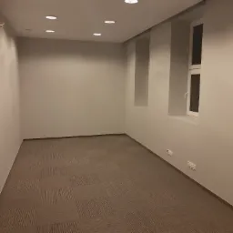 Pomalowane na szaro wnętrze biura z wykładziną dywanową w kwadraty, widoczne okno i gniazdka elektryczne.