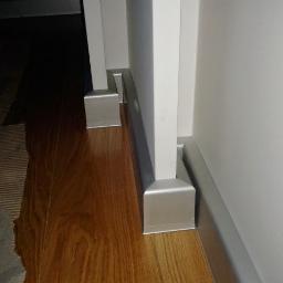 Paweł Łuczak Usługi remontowo budowlane - Szczegółowe ujęcie nowo położonej, błyszczącej, drewnianej podłogi z przypodłogowymi listwami w kolorze srebrnym, kontrastującymi z białą ścianą i framugą drzwi.