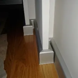 Szczegółowe ujęcie nowo położonej, błyszczącej, drewnianej podłogi z przypodłogowymi listwami w kolorze srebrnym, kontrastującymi z białą ścianą i framugą drzwi.