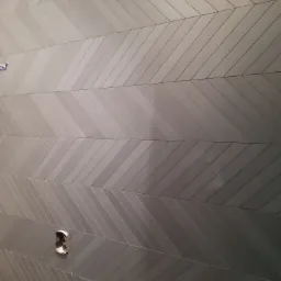 Ściana wyłożona szarymi, błyszczącymi płytkami ułożonymi we wzór jodełki, z widocznym otworem na gniazdko elektryczne w dolnej części.