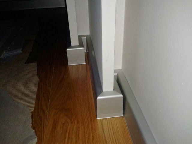 Szczegółowe ujęcie nowo położonej, błyszczącej, drewnianej podłogi z przypodłogowymi listwami w kolorze srebrnym, kontrastującymi z białą ścianą i framugą drzwi.