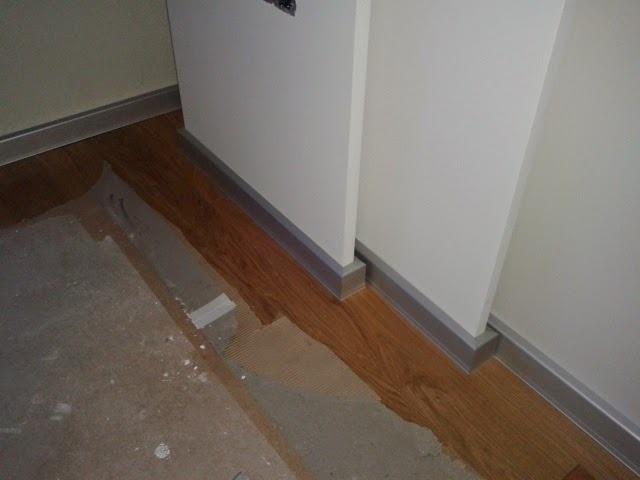 Fragment podłogi z panelami drewnopodobnymi i szarą listwą przypodłogową, widoczne ślady prac remontowych i pozostałości kleju na podłożu.