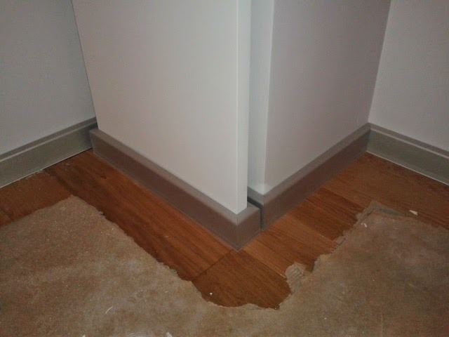 Detal podłogi z drewnianą podłogą i szarą listwą przypodłogową, widoczne fragmenty starej wykładziny.