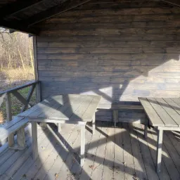 Drewniana altana z dwoma stołami i ławką, pomalowana na szaro, z widokiem na drzewa bez liści i resztki śniegu na ziemi.