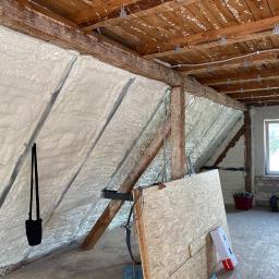 FUH Met-Pio Piotr Kuboszek - Poddasze w trakcie ocieplania pianką poliuretanową, widoczne drewniane belki konstrukcyjne, fragment okna oraz elementy wyposażenia budowlanego, takie jak płyta OSB i narzędzia.