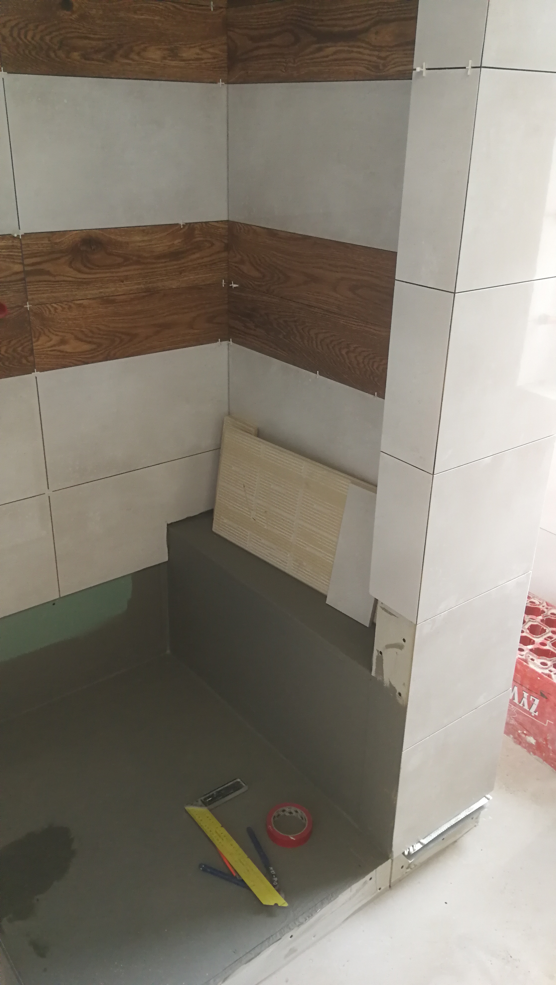 Wnętrze prysznica w trakcie kładzenia płytek: szare i drewnopodobne płytki ułożone naprzemiennie na ścianach, widoczne narzędzia glazurnicze, folia w płynie na podłodze.
