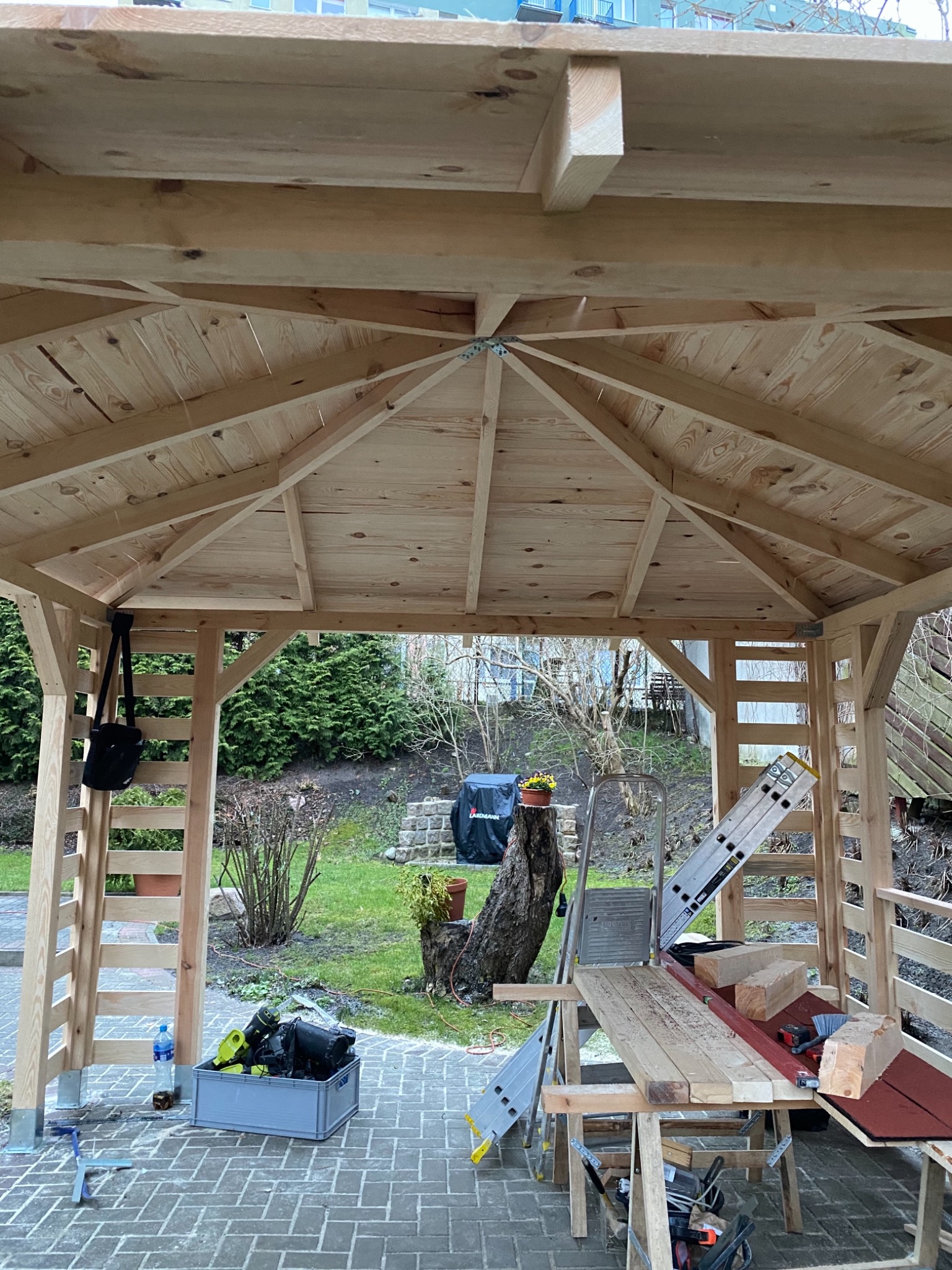 Wnętrze w trakcie budowy drewnianej altany ogrodowej, widoczne elementy konstrukcyjne dachu i ścian, drabina, stół roboczy z narzędziami i materiałami budowlanymi, otoczenie zieleni ogrodowej.