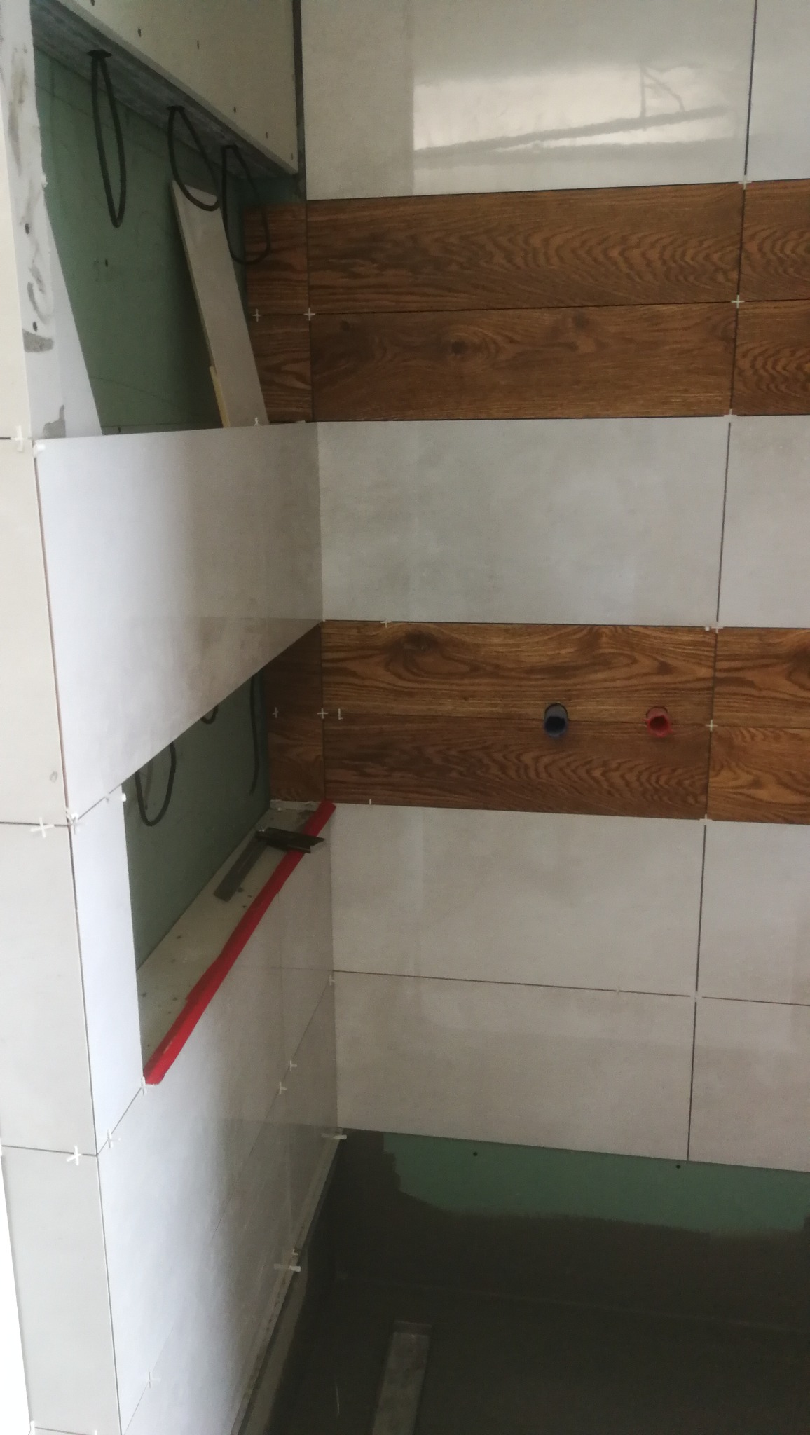 Montaż płytek w łazience w Olsztynie: białe płytki ścienne i dekoracyjne panele drewnopodobne, widoczne krzyżyki dystansowe, instalacja hydrauliczna w ścianie, etap prac wykończeniowych.